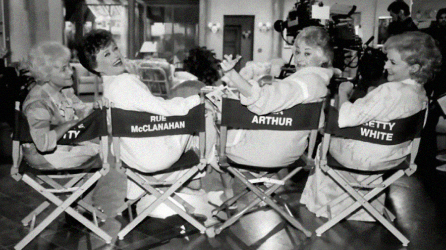 Four women sit in director’s chairs labeled with their names on a TV set, smiling and looking back at the camera. Crew and equipment are visible in the background. The image is black and white.