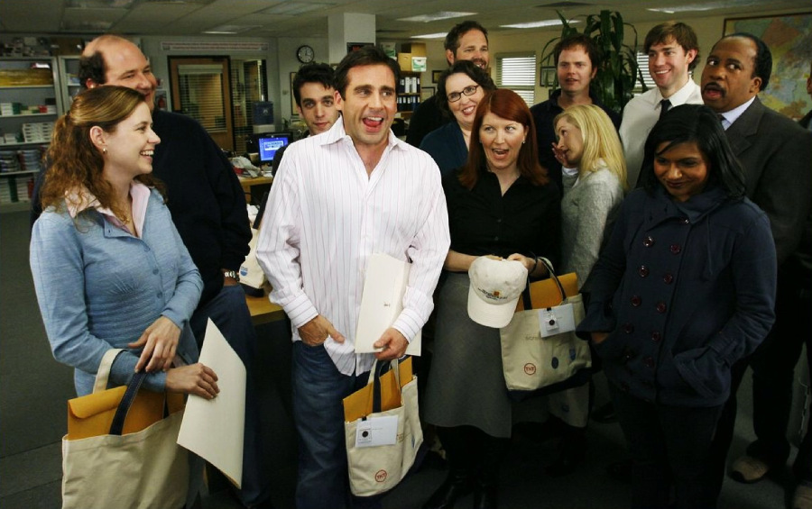 A group of twelve office workers stand together in an office, smiling and laughing. Some hold tote bags and envelopes, and the setting appears friendly and informal with desks and office supplies in the background.