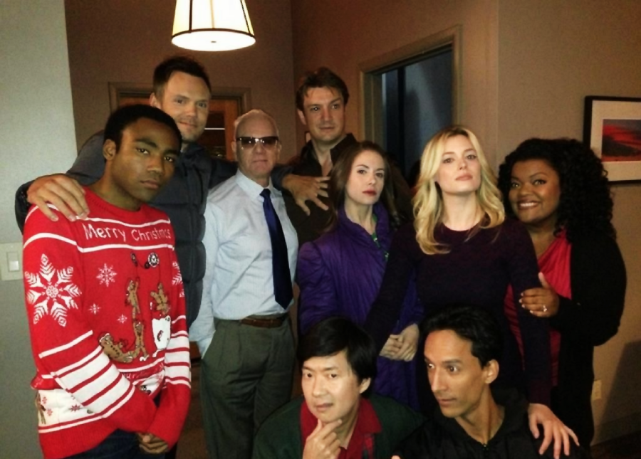 Nine people pose together indoors. One person wears a festive red Christmas sweater, while others are dressed casually or in dark colors. They stand and kneel closely, some smiling, in a warmly lit room with beige walls and a hanging lamp.