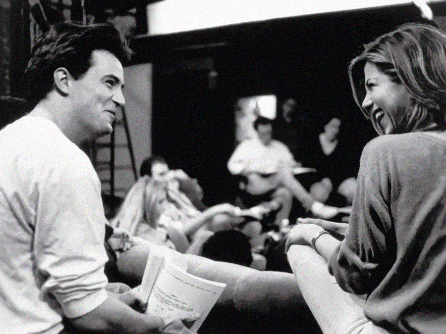 Two people sit close together smiling and talking, holding scripts, with others in the background. The setting appears relaxed and casual, possibly backstage or during a rehearsal. The photo is in black and white.