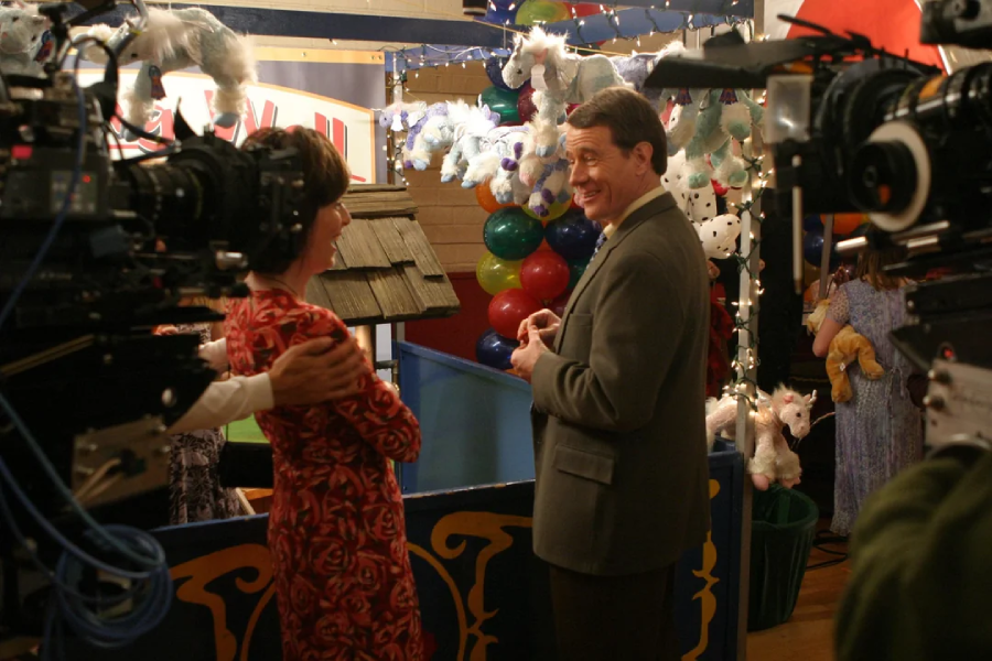 Two actors stand smiling and talking at a carnival booth decorated with plush unicorns and colorful balloons, while cameras and crew film the scene around them.