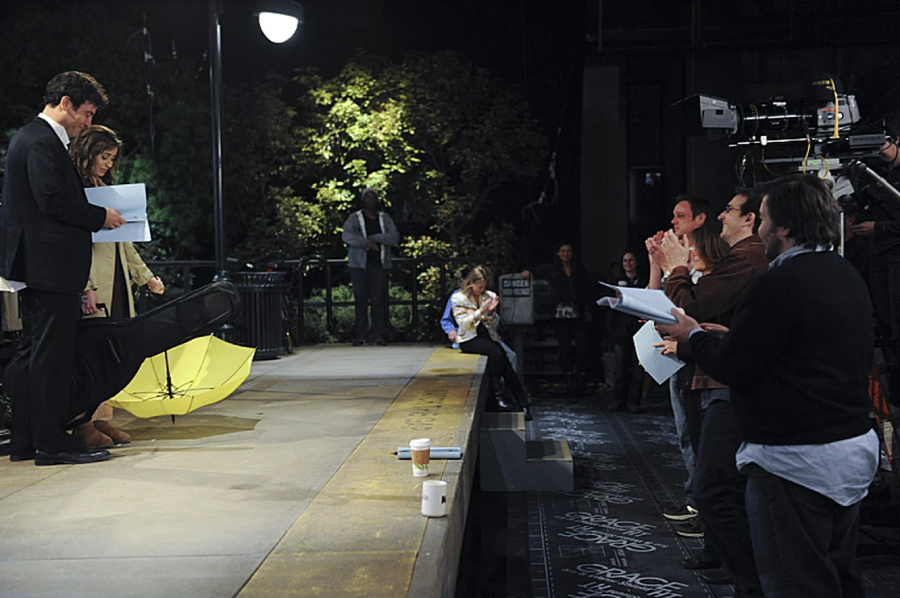 Actors stand on an outdoor set holding scripts near a yellow umbrella, while crew members with cameras and scripts watch and clap. Coffee cups and equipment are on the ledge, and people are gathered in the background.