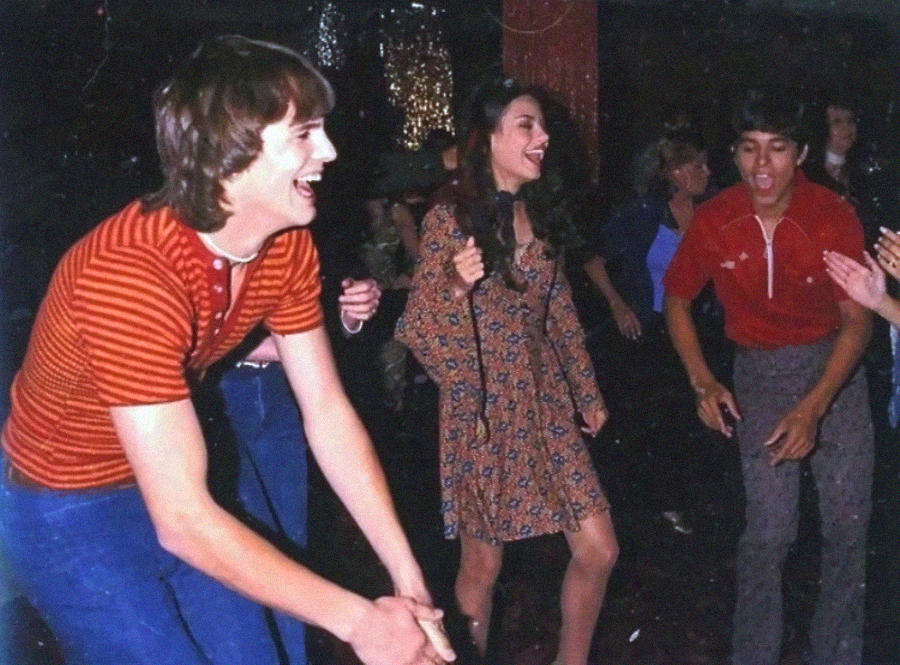 Three young people are laughing and dancing together at a lively party, surrounded by others. The scene is energetic, with festive clothing and dim, colorful lighting in the background.