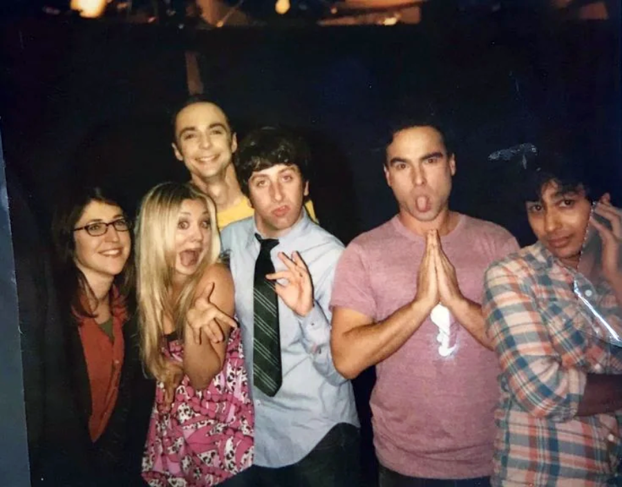 Six people stand close together, making silly faces and gestures for the camera, in what appears to be a behind-the-scenes or casual setting with dark background and warm lighting.