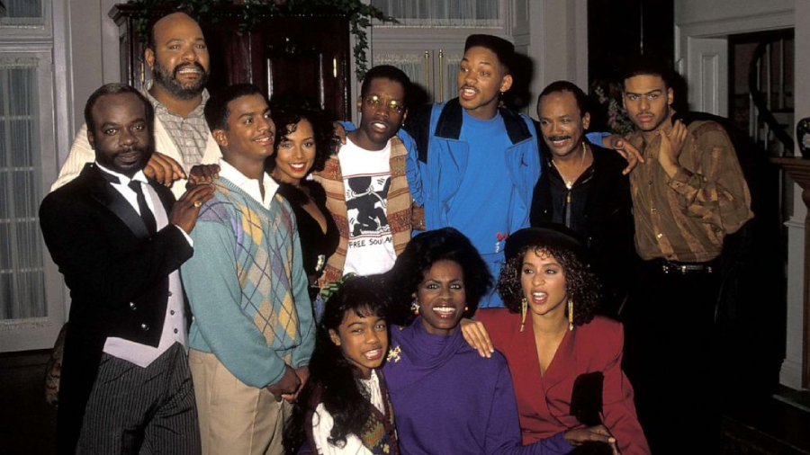 A group of twelve people, smiling and posing together indoors. They are dressed in 1990s-style clothing, and some have their arms around each other, creating a sense of camaraderie and happiness.