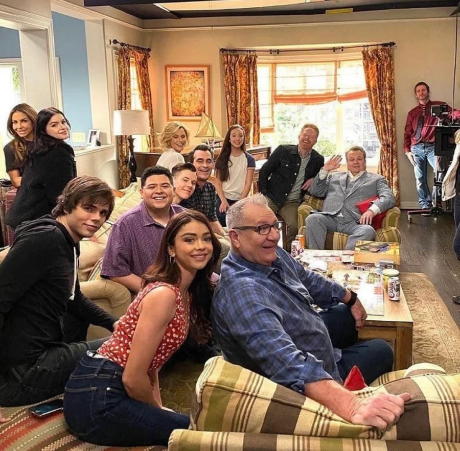 A group of people, some sitting on couches and chairs and others standing behind them, smile and pose for a photo in a cozy, brightly lit living room set.