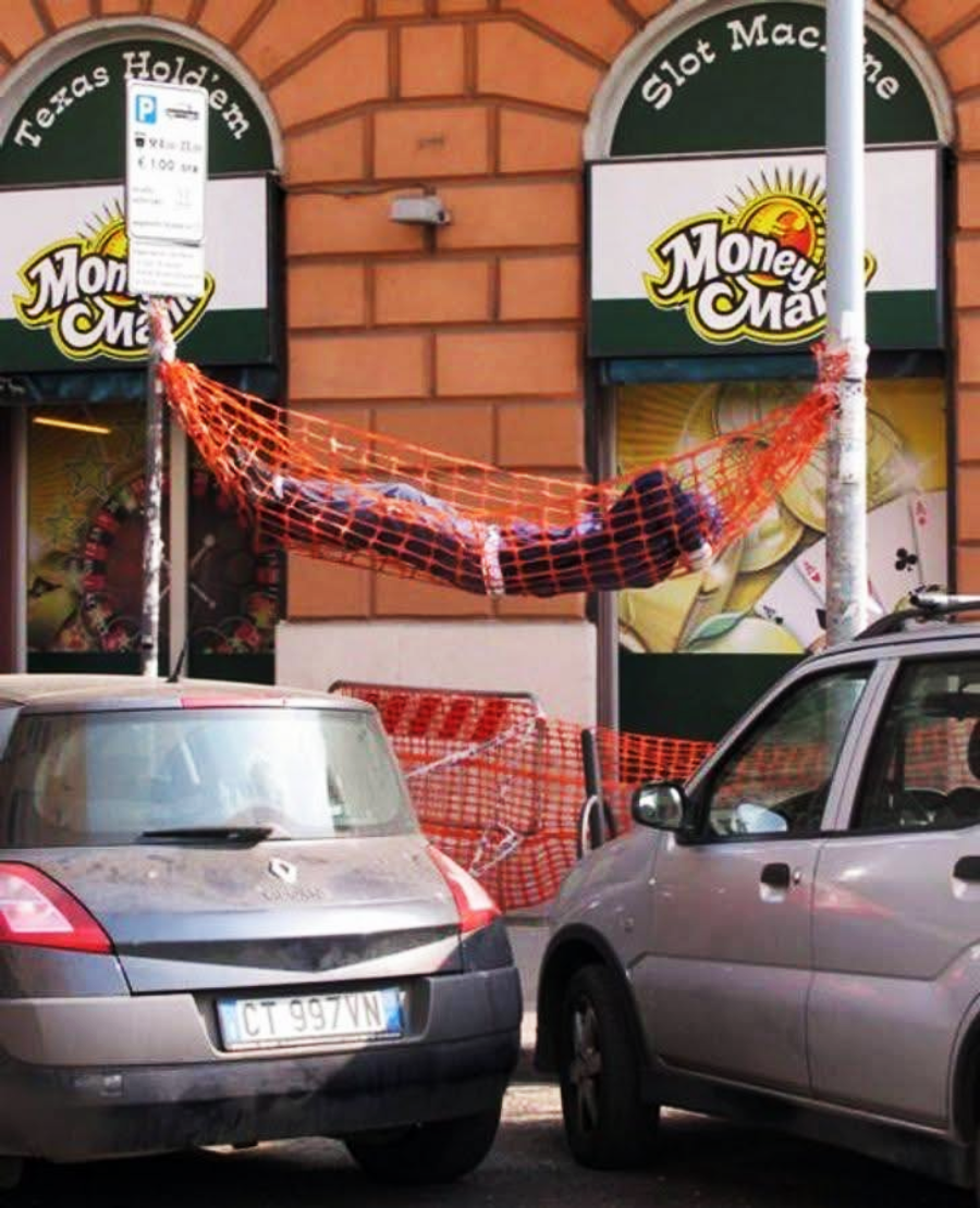 A person is lying in an orange construction net hammock strung between two poles on a city street, with parked cars and a "Money Mart" storefront in the background.