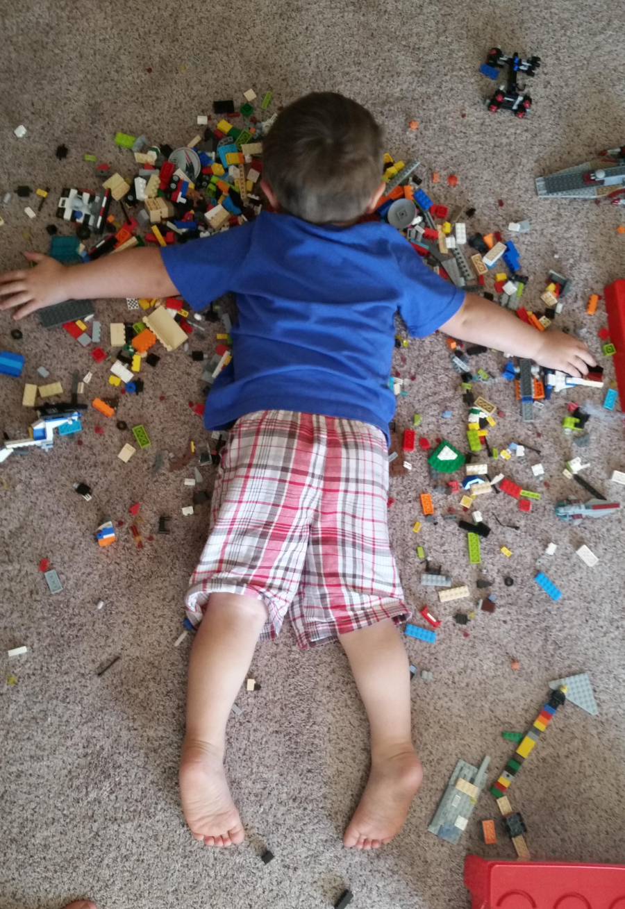 A young child wearing a blue shirt and plaid shorts lies face down on a carpet surrounded by scattered colorful LEGO bricks and pieces.