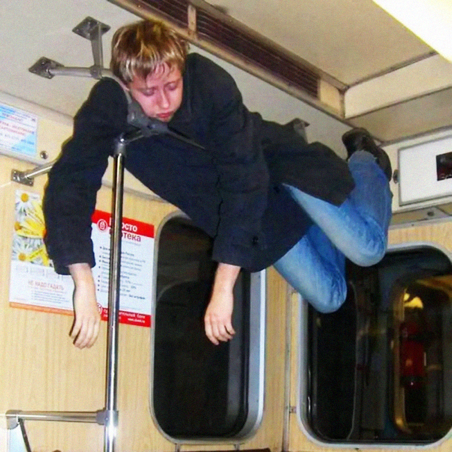 A person in a dark coat and jeans is sleeping awkwardly while hanging horizontally between a subway pole and the wall inside a train carriage.