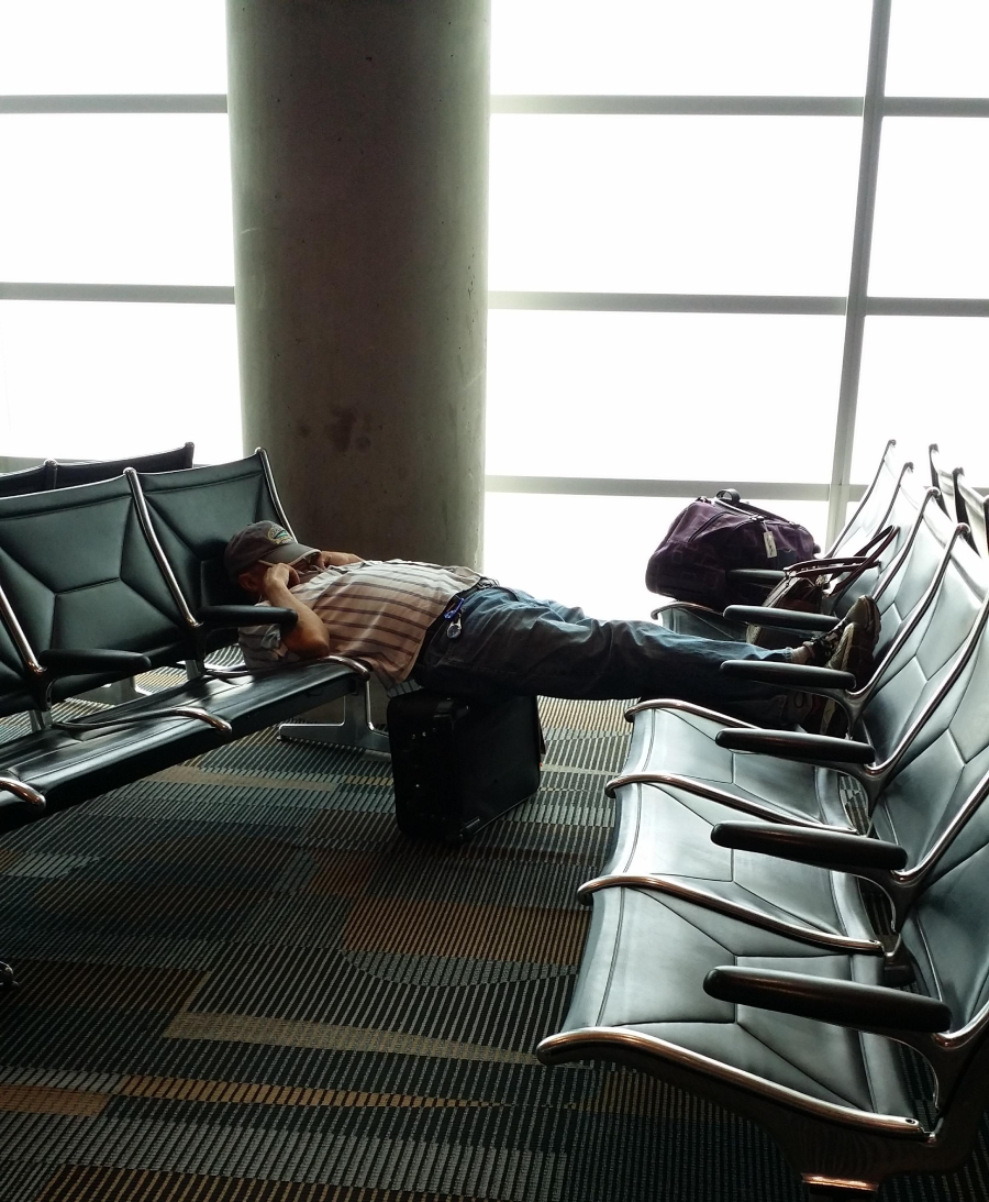 A person lies on airport seating with a hat covering their face, legs stretched across seats and feet resting on a suitcase. A backpack is placed on a nearby chair by a large window and concrete pillar.