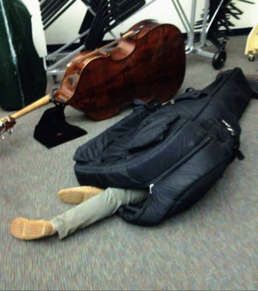 A person is lying on the floor with their legs sticking out of a large black instrument case, with a cello and other cases visible in the background, suggesting a music room setting.