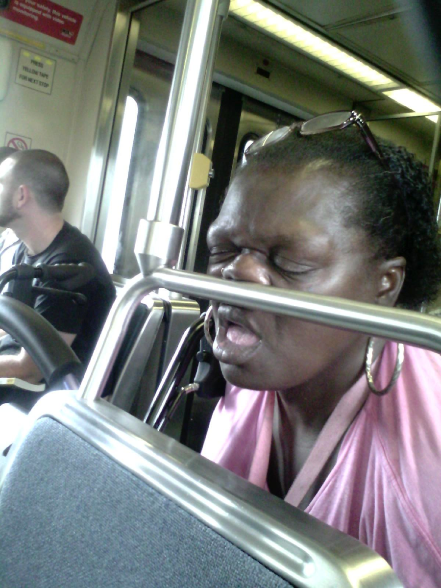 A woman wearing glasses on her head and large hoop earrings sleeps upright on public transit, her face pressed against a metal rail. Other passengers are visible in the background.