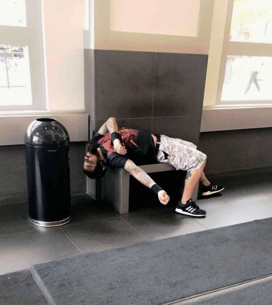 A person with tattoos lies awkwardly and sprawled backward on a bench in a public indoor space, near a large trash can and windows, appearing to rest or sleep.