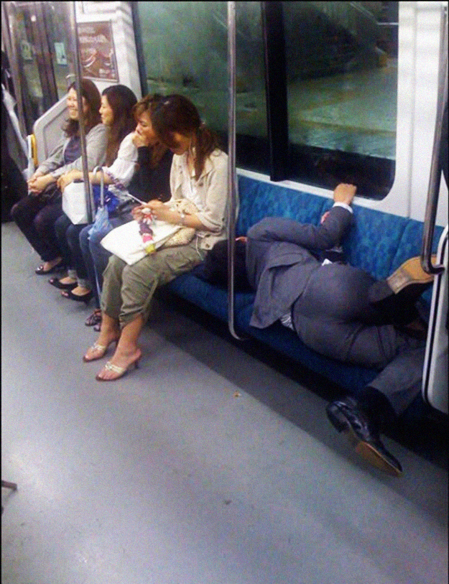 Four women sit on a subway bench while a man in a suit lies down, taking up several seats with his legs stretched out and his head resting against the window. The women appear calm and are looking at their phones.