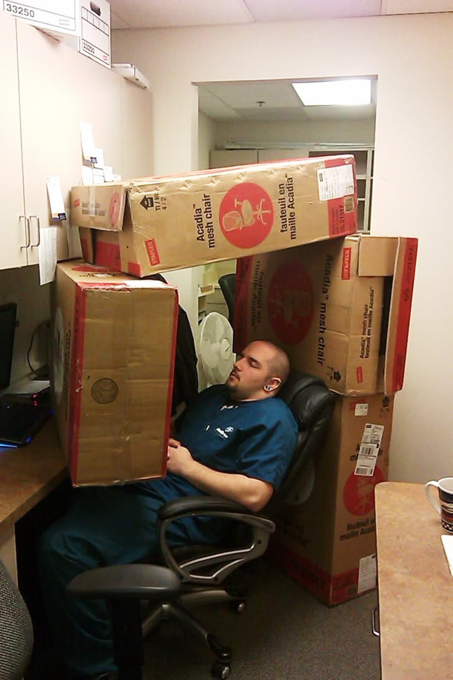 A man in scrubs sleeps in an office chair, surrounded and partially covered by large cardboard boxes stacked around him at his desk.