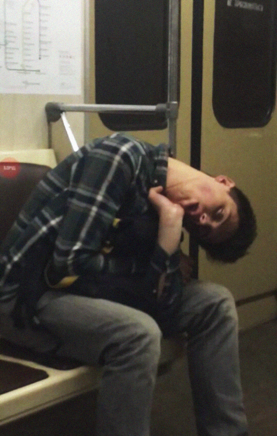 A person wearing a plaid shirt and jeans is slumped over and sleeping awkwardly on a subway seat, with their head tilted back and mouth open. A subway map is visible on the wall behind them.