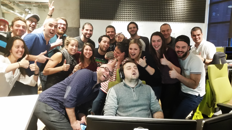 A group of cheerful people crowd around a man sleeping in a chair at his desk, smiling, laughing, and giving thumbs up in an office setting.