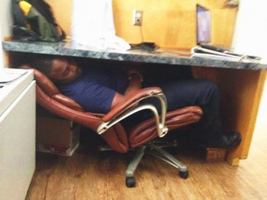 A person is lying back and sleeping in an office chair that is reclined far under a desk, with their feet sticking out and their arms crossed. A laptop and other office items are on the desk above them.
