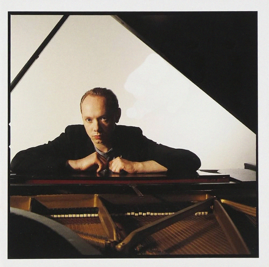 A person sits behind a grand piano, resting their arms on the edge and looking directly at the camera. The piano strings and lid are visible in the foreground. The background is plain and well-lit.