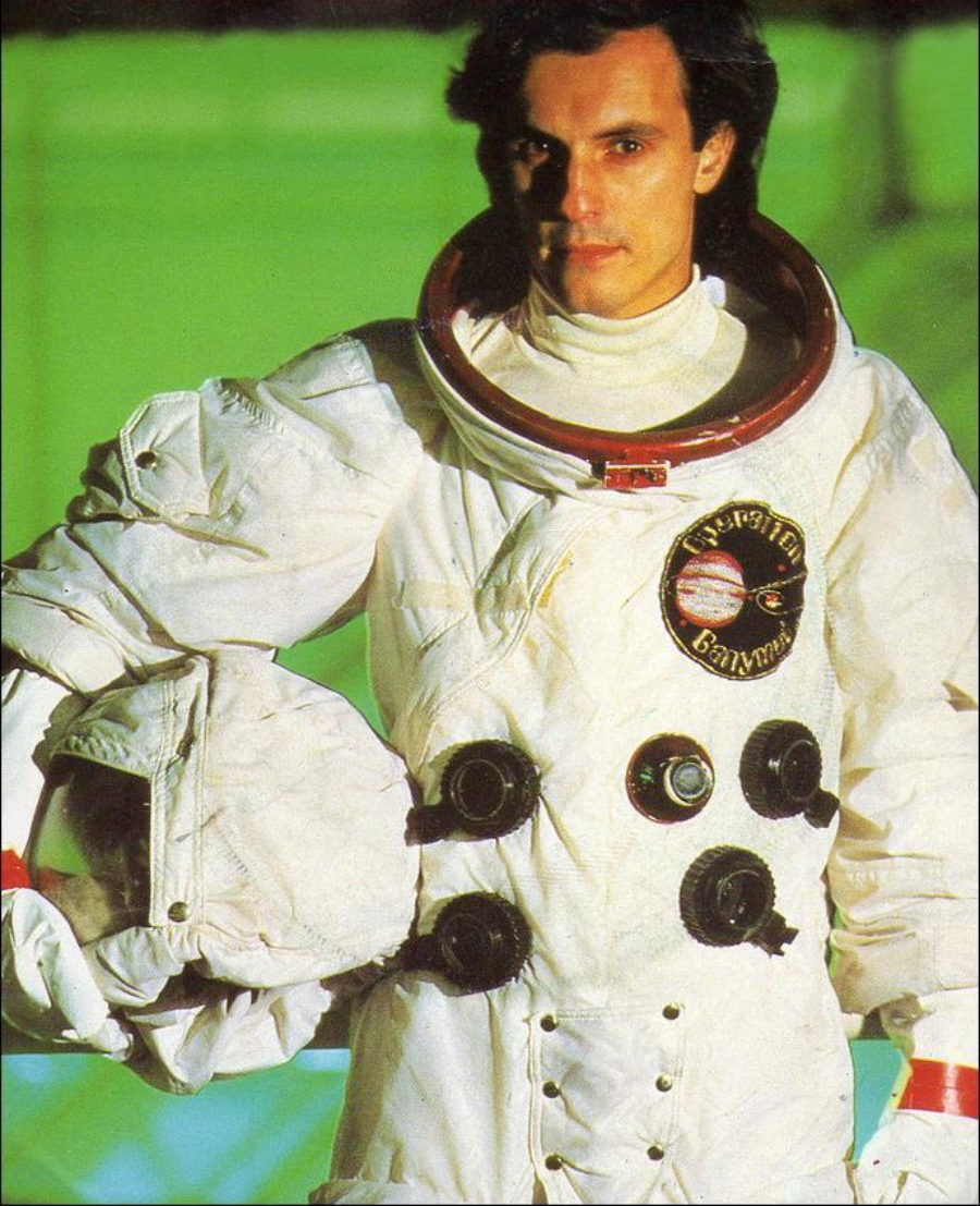 A person wearing a white spacesuit with a helmet tucked under their arm stands in front of a green background. The suit has several circular valves and a patch featuring Jupiter and the word "Discovery.