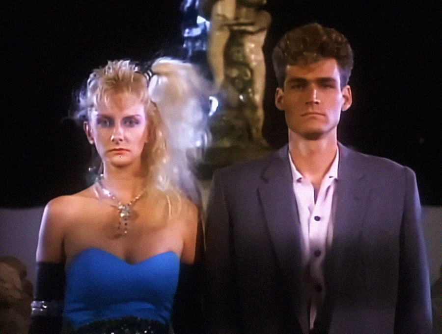 A woman in a blue strapless dress with blonde hair stands next to a man in a gray suit and white shirt. Both have serious expressions. A fountain or statue is visible in the dimly lit background.
