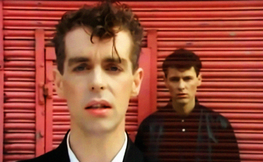 Two men stand in front of a closed, red metal shutter. The man in the foreground has curly hair and wears a dark jacket, while the man in the background stands slightly out of focus, also dressed in dark clothing.