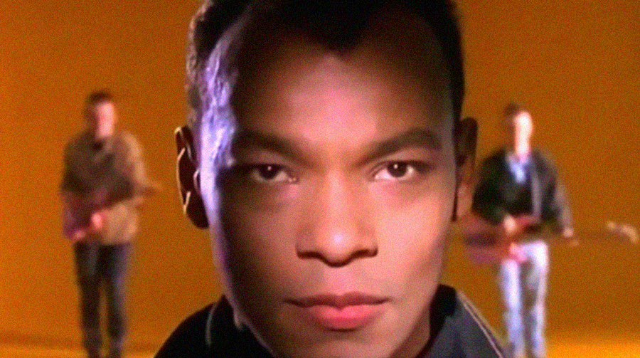 A close-up of a man's face in sharp focus, with two blurry figures holding guitars in the orange-lit background.