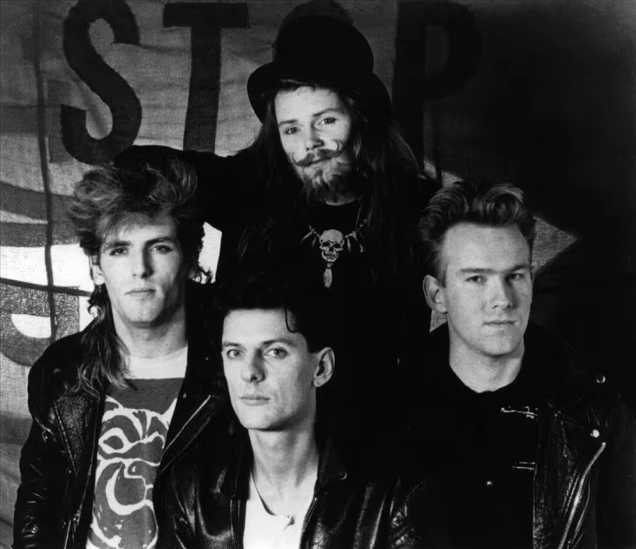 Four men pose together in front of a backdrop with the word “STOP.” Three are wearing leather jackets, and one at the back wears a hat and a beard. The image is black and white and has a retro, rock band style.