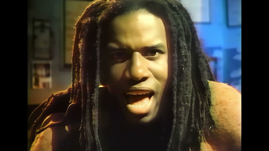 A person with long, dark dreadlocks and an intense expression is looking directly at the camera, mouth open as if speaking or singing, with a blurred background.