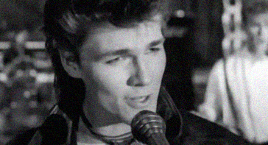 A black-and-white close-up of a young man singing into a microphone, with a focused expression. The background is blurred and features another person and some equipment.