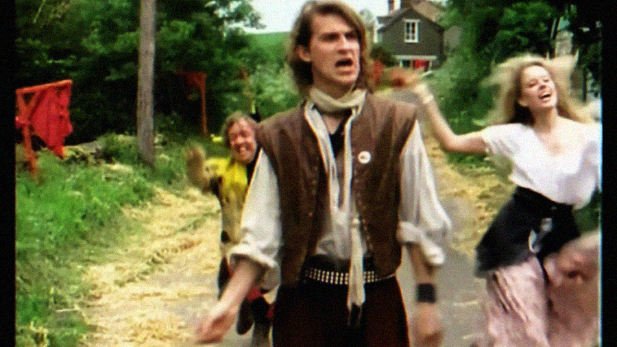 Three people in colorful, old-fashioned clothing are joyfully running down a country road with trees and a house in the background. The scene feels energetic and playful.