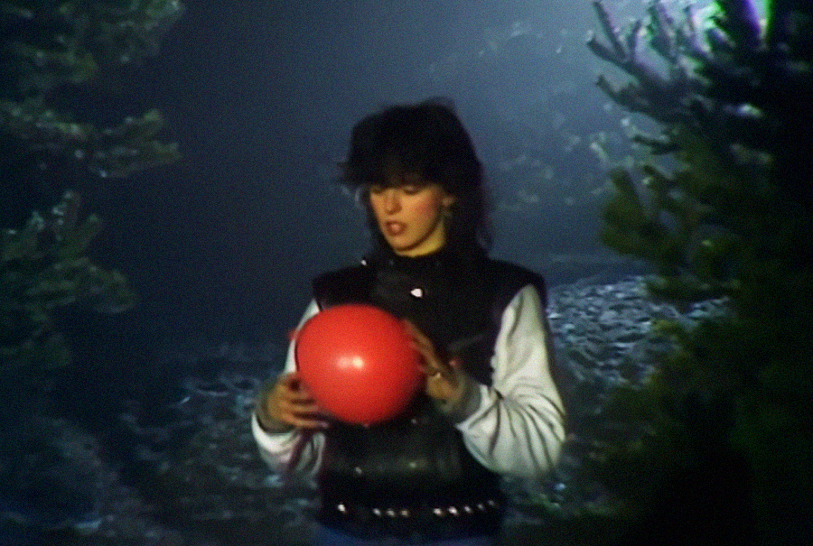 A person with dark hair, wearing a black vest and white sleeves, stands outdoors at night, holding a red balloon between two trees. Moonlight or artificial light illuminates the scene.