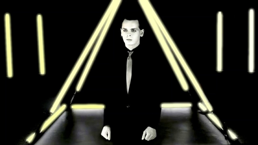 A person in a suit and tie stands in front of a glowing triangular structure, surrounded by vertical and horizontal yellow lights in a dark, futuristic setting.