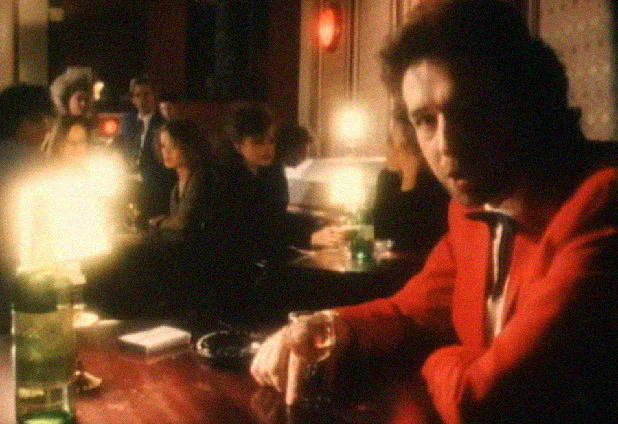 A man in a red jacket sits at a dimly lit bar holding a glass, with other people gathered and talking in the blurred background. Warm lighting gives the scene a soft, nostalgic atmosphere.