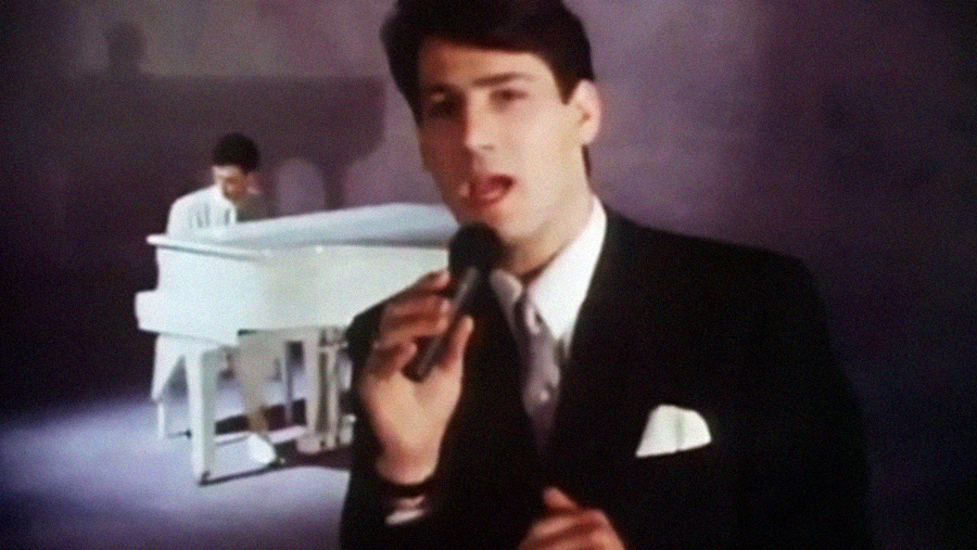 A man in a dark suit holds a microphone and sings, while another person plays a white grand piano in the background on a softly lit stage.