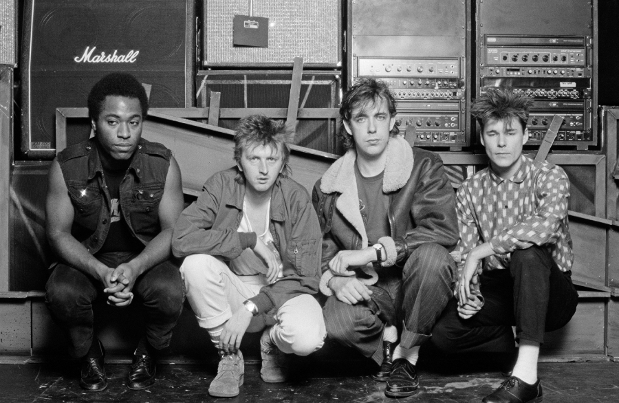 Four men sit crouched together in front of stacked amplifiers and audio equipment. They wear casual, 1980s-style clothing and look directly at the camera with serious expressions.