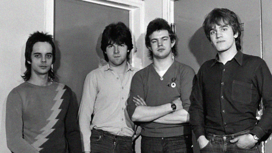 Four young men stand indoors in front of a door, posing for a black-and-white photo. They wear casual clothes and have various hairstyles typical of the late 1970s or early 1980s.