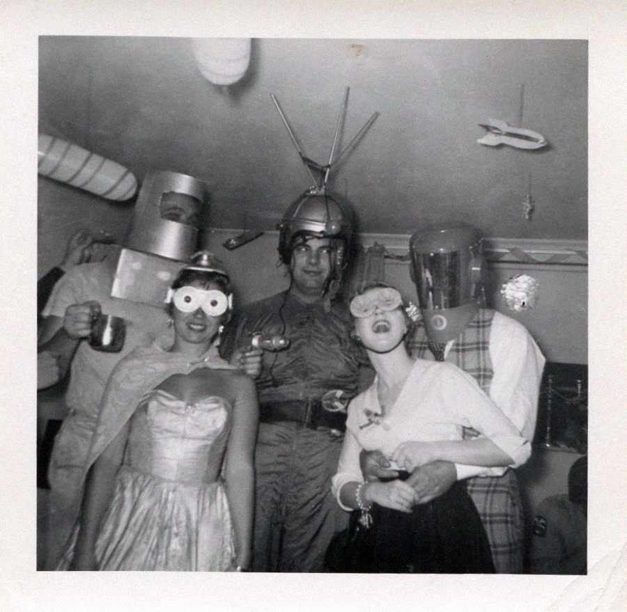 Six people at a costume party wear creative outfits, including robot helmets, goggles, and a space suit with an antenna. Two women stand in front wearing dresses and novelty glasses. The background has quirky decorations.