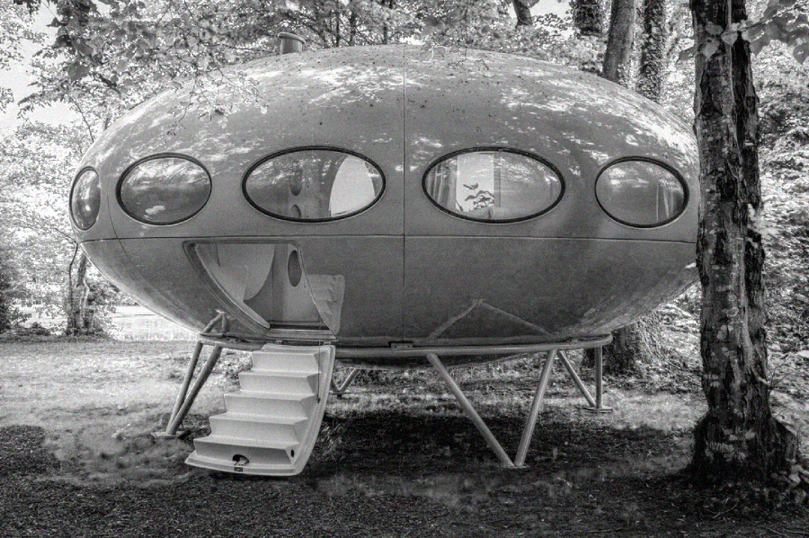 A round, spaceship-like house with oval windows and a staircase leading to an open doorway, set among trees in a wooded area.