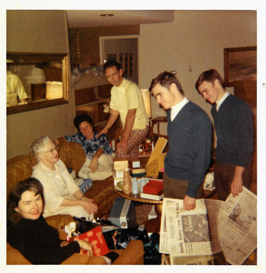 A vintage photo of six adults, four seated and two standing, gathered in a living room with gifts, boxes, and newspapers spread out, suggesting a holiday or special occasion. The setting appears warm and casual.