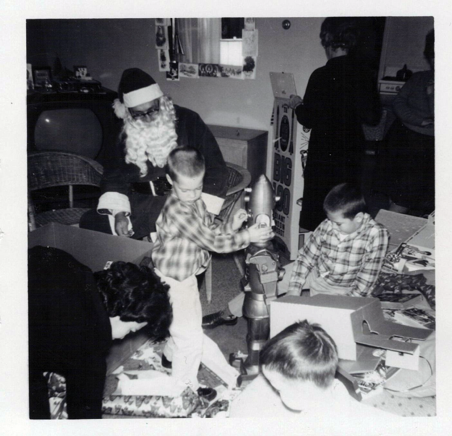 Black and white photo of children opening presents near a person dressed as Santa Claus. Two boys are playing with a rocket toy; an adult bends over wrapping paper, and others stand in the background.