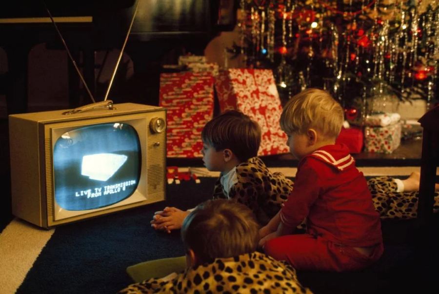 Three young children in pajamas sit on the floor watching an old-fashioned television. Behind them, a decorated Christmas tree and wrapped presents are visible, creating a festive holiday scene.