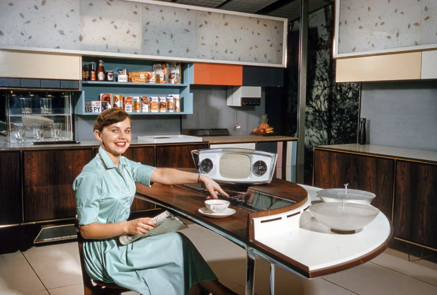 A woman in a retro-futuristic kitchen sits at a curved counter with a built-in TV, control panel, and dishes. Shelves with food and appliances line the walls, evoking a 1950s vision of the future.