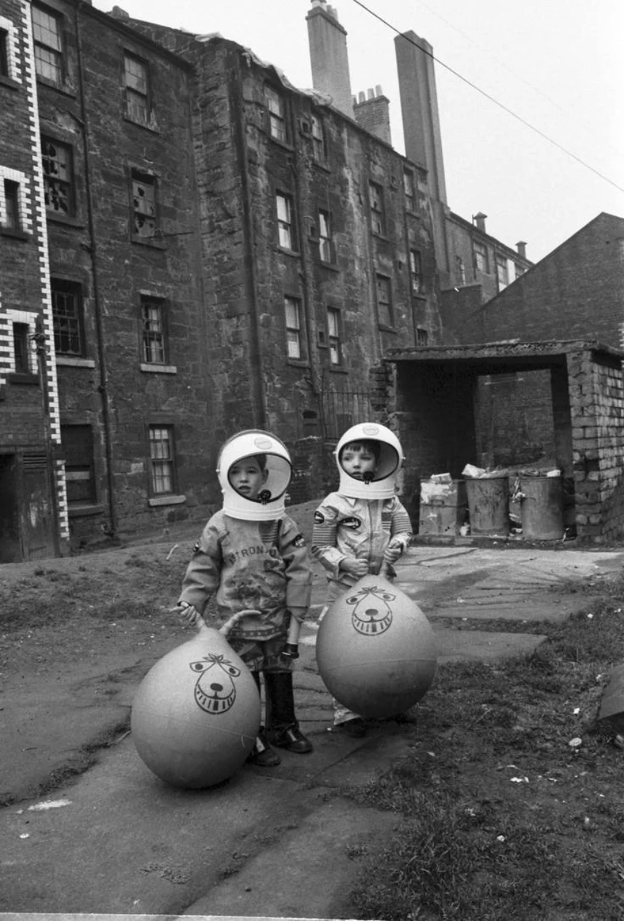 Two children wearing toy space helmets stand outside in an urban courtyard, each holding a large bouncy ball with a smiling face design. Behind them are old brick tenement buildings and a rundown yard.
