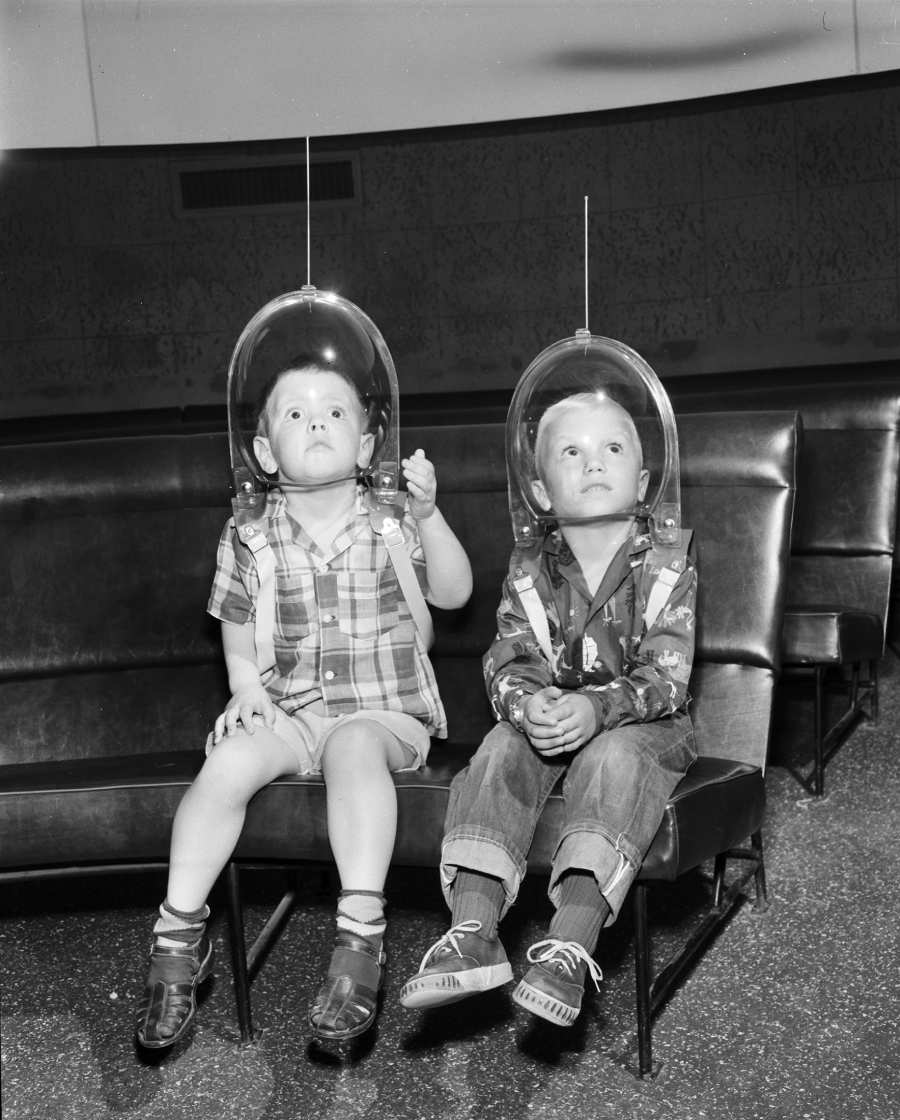Two young children sit on a bench wearing toy space helmets and backpacks, looking upward with interest. One child points while both appear fascinated, suggesting they are imagining being astronauts or exploring space.