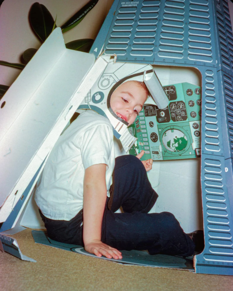 A young child sits inside a cardboard space capsule, wearing a paper helmet and smiling while making a peace sign, with detailed control panels depicted around them.