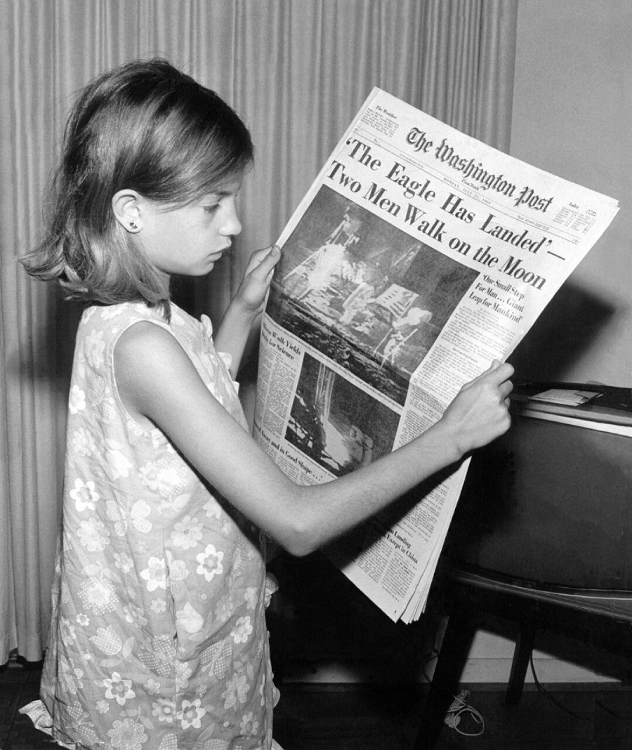 A young girl in a sleeveless floral dress reads a newspaper with the headline "The Eagle Has Landed—Two Men Walk on the Moon" and a photo of astronauts on the lunar surface.