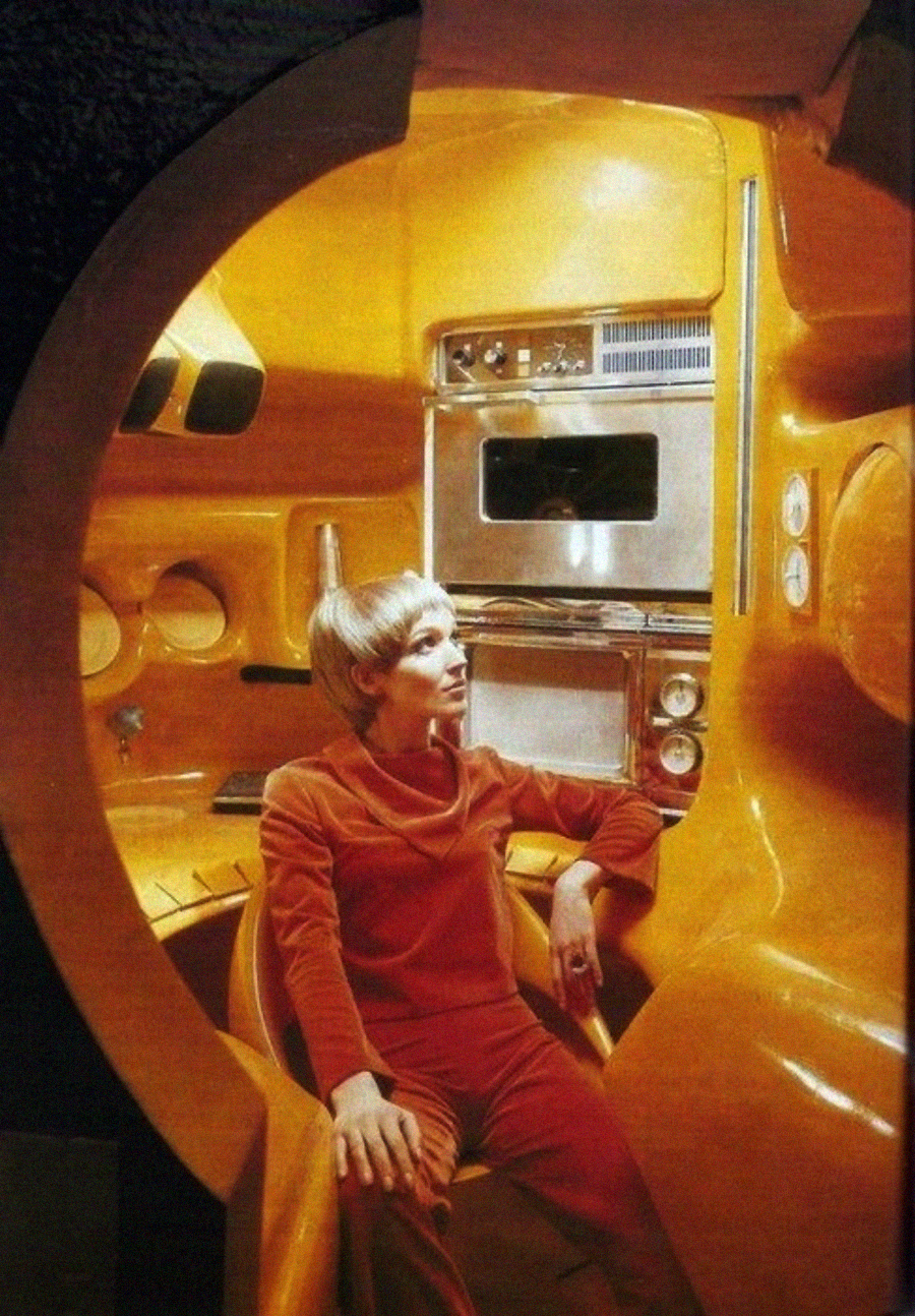 A person in a red outfit sits in a futuristic, rounded orange room with built-in control panels, monitors, and dials, giving a retro science fiction vibe.