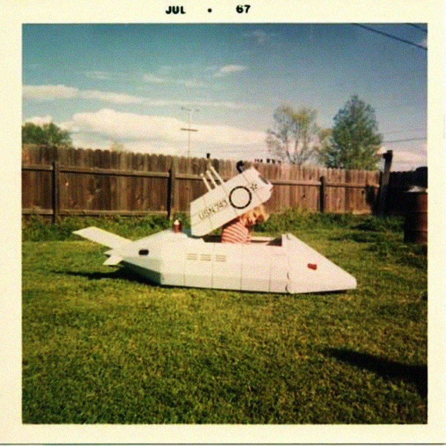 A child sits inside a homemade, toy submarine made of painted cardboard in a grassy backyard, with a wooden fence and trees in the background. The photo is dated July 1967.