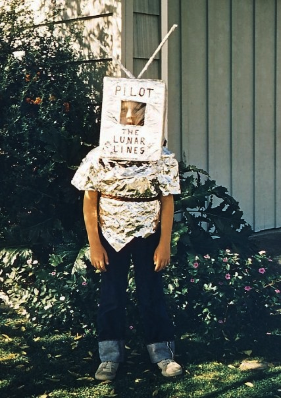 A child stands outside wearing a homemade astronaut costume made of aluminum foil and a box labeled "PILOT THE LUNAR LINES," with an antenna on top. Shrubs and flowers are visible in the background.